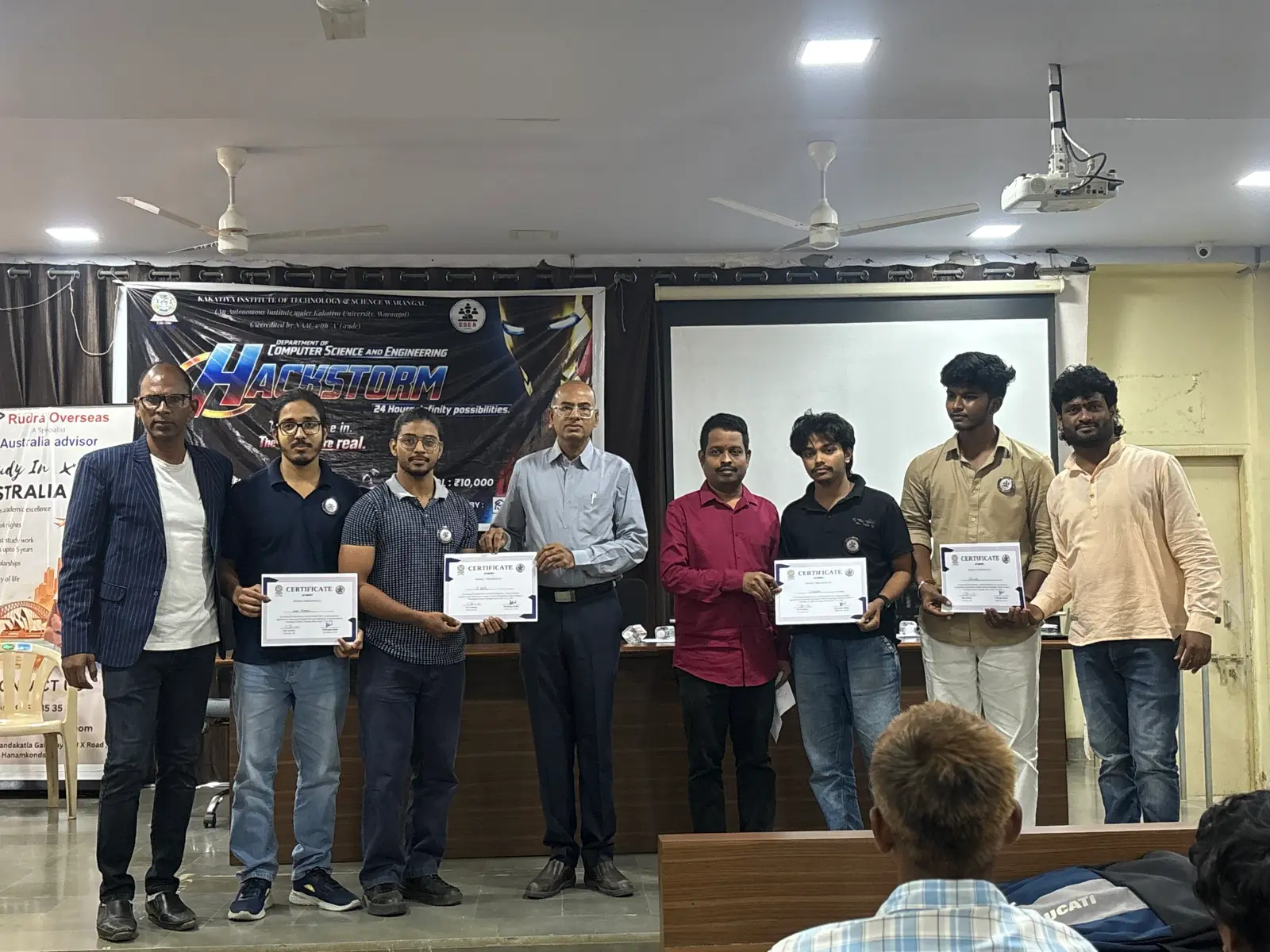 Two winning teams stand with department members, holding certificates in front of the Hackstorm 2026 event banner.