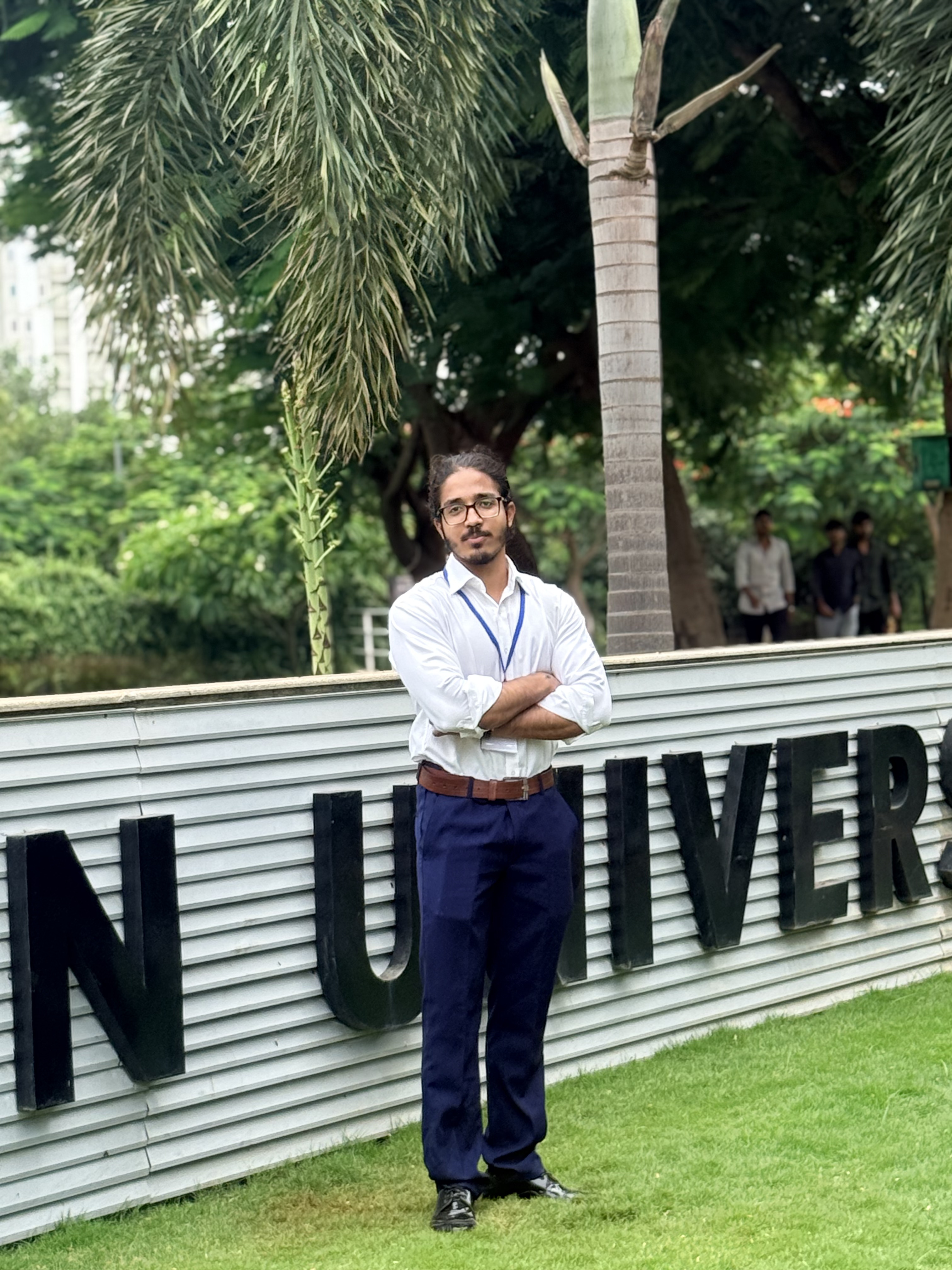 Umair Hazrami standing outdoors in a white shirt, framed by trees and a large sign.